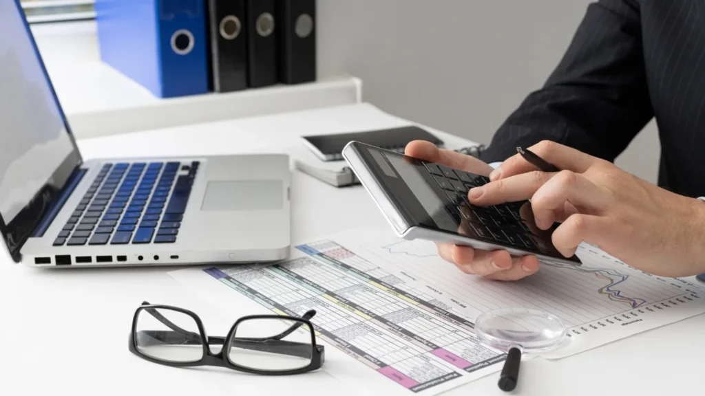 Persona utilizando una tablet y una laptop en un escritorio, con documentos y gafas, ilustrando un ambiente de trabajo moderno y productivo.
